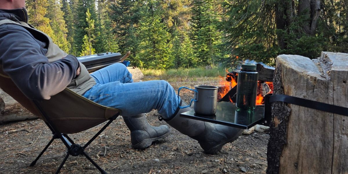 Top Shelf outdoor and camping table mounted to a tree stump in a campsite next to a fire pit holding a coffee cup and water bottle.