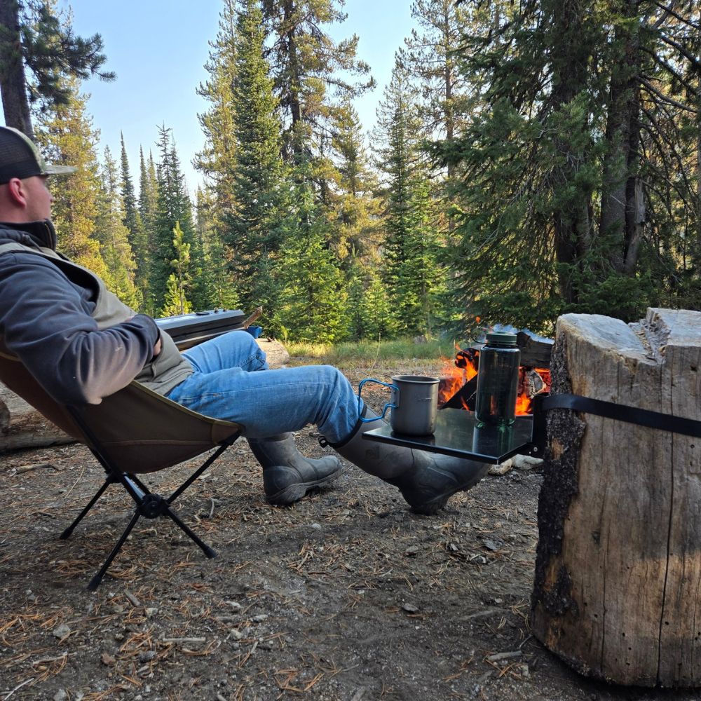 Top Shelf outdoor and camping table mounted to a tree stump in a campsite next to a fire pit holding a coffee cup and water bottle.